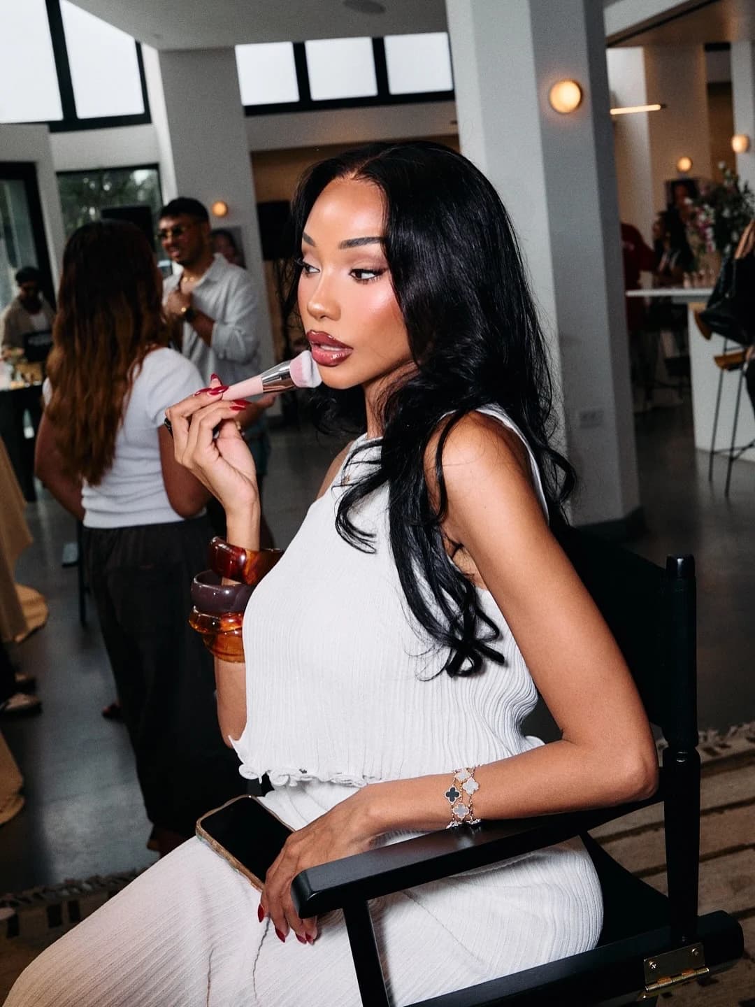 Melly Sanchez applying blush with a brush, seated in a director’s chair at a beauty event